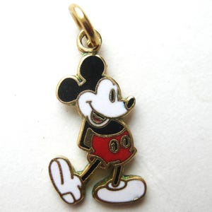 Puede incluir: Un charm de Mickey Mouse dorado con orejas negras, cara blanca y pantalones cortos rojos. El charm tiene un peque&ntilde;o lazo para sujetarlo a un collar o pulsera. El personaje de dibujos animados cl&aacute;sico se representa caminando.