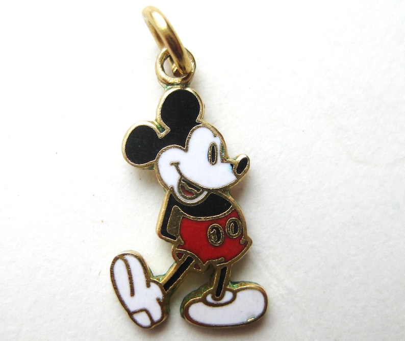 Puede incluir: Un charm de Mickey Mouse dorado con orejas negras, cara blanca y pantalones cortos rojos. El charm tiene un peque&ntilde;o lazo para sujetarlo a un collar o pulsera. El personaje de dibujos animados cl&aacute;sico se representa caminando.