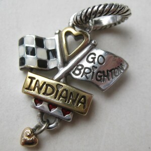 May include: A silver charm with a checkered flag, a flag that says "GO BRIGHTON", and a banner that says "INDIANA". The charm has a gold heart and red heart accents. The charm has a silver twisted ring at the top.