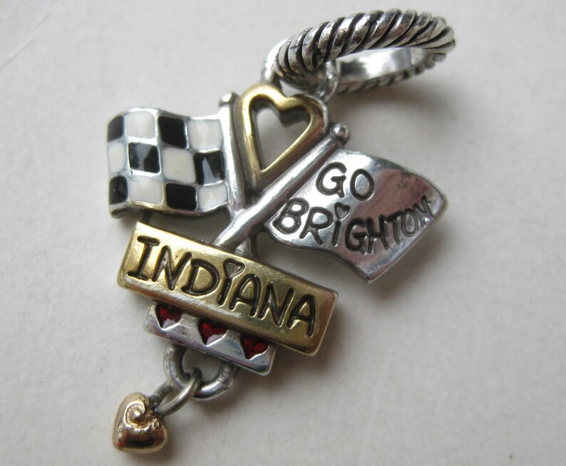 May include: A silver charm with a checkered flag, a flag that says "GO BRIGHTON", and a banner that says "INDIANA". The charm has a gold heart and red heart accents. The charm has a silver twisted ring at the top.