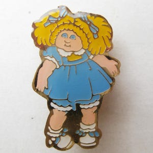 May include: A metal enamel pin featuring a cartoon doll with bright yellow hair in pigtails, wearing a blue dress with a white collar, blue bloomers, and white shoes with blue accents. The doll has light skin, blue eyes, and a small nose.