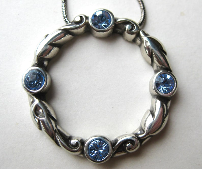 May include: A silver-toned pendant necklace featuring a circular design with five light blue gemstones. The pendant has a detailed, leaf-like pattern around the gemstones. The necklace chain is a thin, silver-colored snake chain.