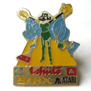 Könnte beinhalten: Vintage Atari Lynx Anstecknadel mit einer stilisierten Figur in grünem Outfit mit ausgestreckten Armen, vor gelbem Hintergrund. Die Nadel enthält den Text "Ishido" und "ATARI" in Schwarz, mit einem roten Dreieck und blauen Akzenten.