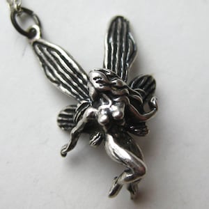 Peut inclure: Un collier pendentif argenté représentant une fée détaillée. La fée a des ailes déployées et des cheveux flottants, avec des détails complexes sur les ailes et le corps. Le pendentif est suspendu à une fine chaîne argentée.