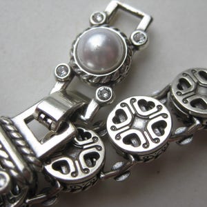Puede incluir: Una pulsera de tono plateado con eslabones circulares alternos con un patr&oacute;n de corazones recortados y eslabones redondos engastados con una perla blanca central y peque&ntilde;as piedras claras. Los componentes met&aacute;licos tienen una textura trenzada.