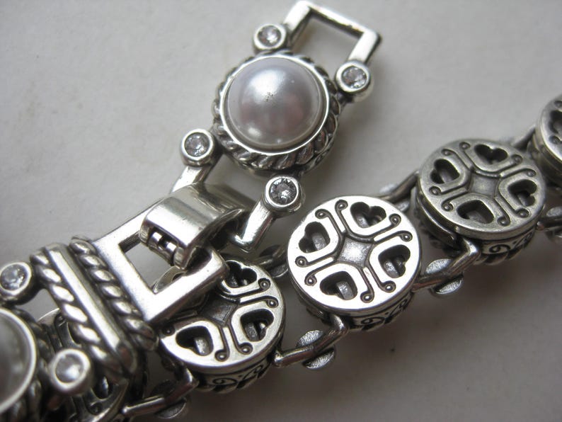 Puede incluir: Una pulsera de tono plateado con eslabones circulares alternos con un patr&oacute;n de corazones recortados y eslabones redondos engastados con una perla blanca central y peque&ntilde;as piedras claras. Los componentes met&aacute;licos tienen una textura trenzada.