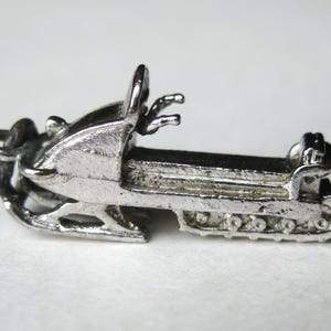 Może przedstawiać: Mały, srebrny charms w kształcie skutera śnieżnego. Charms przedstawia szczeg&oacute;łowy projekt skutera śnieżnego z siedzeniem, kierownicą i gąsienicami. Charms jest prawdopodobnie wykonany z metalu i ma być wisiorkiem lub zawieszką.