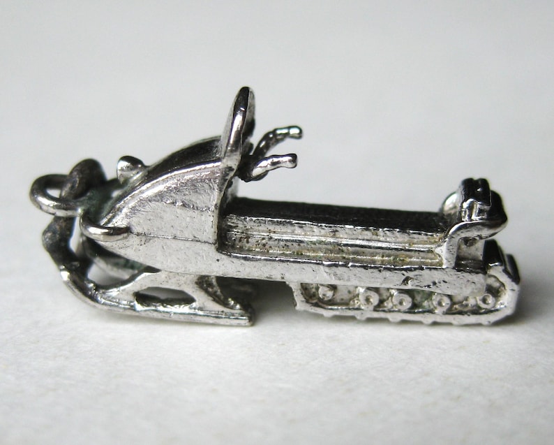 Może przedstawiać: Mały, srebrny charms w kształcie skutera śnieżnego. Charms przedstawia szczeg&oacute;łowy projekt skutera śnieżnego z siedzeniem, kierownicą i gąsienicami. Charms jest prawdopodobnie wykonany z metalu i ma być wisiorkiem lub zawieszką.