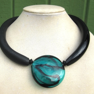 Könnte beinhalten: Eine schwarze Statement-Halskette mit einem großen, türkis-schwarzen Glasanhänger. Die Halskette hat ein dickes, abgerundetes schwarzes Band. Der Anhänger ist ein runder, türkisfarbener Glasanhänger mit schwarzen Wirbeln. Auf einer weißen Schaufensterpuppe präsentiert.