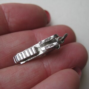 Może przedstawiać: Mały, srebrny wisiorek w kształcie zabytkowego aparatu fotograficznego. Urok ma szczeg&oacute;łowe grzbiety i pętlę do mocowania. Na uroku wytłoczono napis "STERLING".