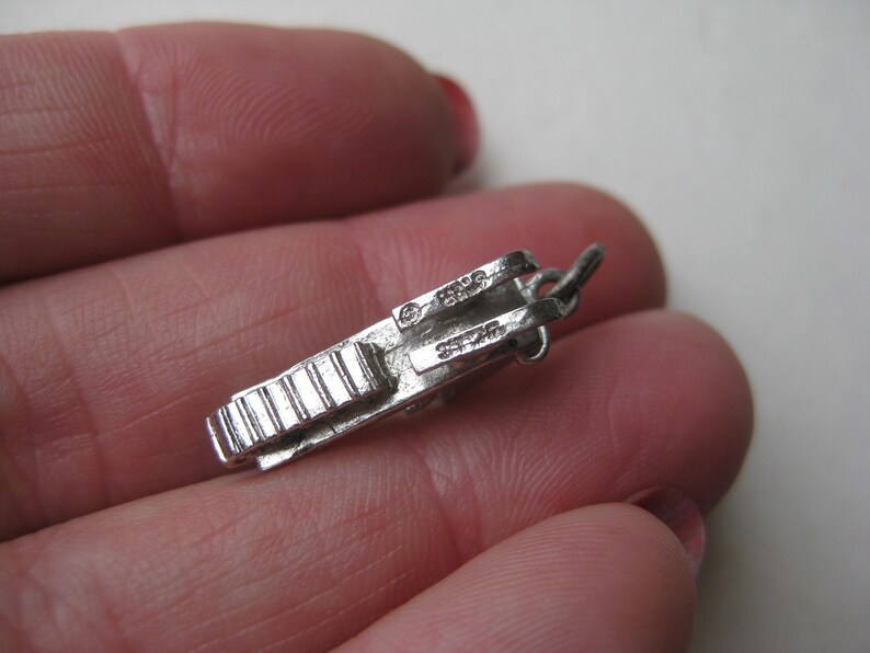 Może przedstawiać: Mały, srebrny wisiorek w kształcie zabytkowego aparatu fotograficznego. Urok ma szczeg&oacute;łowe grzbiety i pętlę do mocowania. Na uroku wytłoczono napis "STERLING".