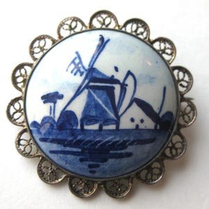 May include: A round brooch featuring a white ceramic center with a blue hand-painted windmill landscape design. The circular ceramic piece is framed by a decorative silver-toned metal border with an intricate, openwork filigree scalloped edge.