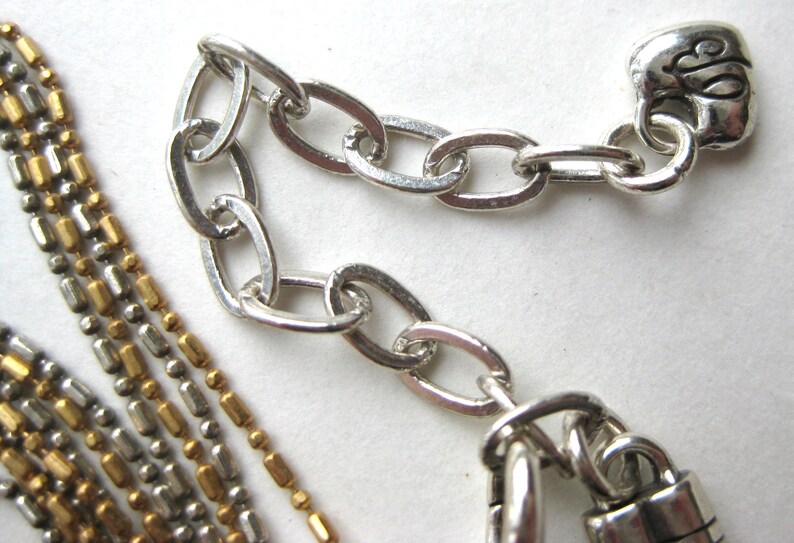 Puede incluir: Un collar de m&uacute;ltiples hilos con cuentas met&aacute;licas que alternan tonos dorados y plateados. El collar incluye una cadena extensora plateada con eslabones ovalados y un dije en forma de coraz&oacute;n al final, con un dise&ntilde;o de letra grabada en cursiva.