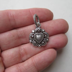 May include: A silver-toned metal pendant featuring a central heart shape surrounded by intricate scrollwork and beaded detailing. The pendant includes a twisted rope-textured bail at the top, showcasing a polished finish with darkened recessed areas.