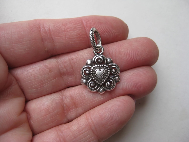May include: A silver-toned metal pendant featuring a central heart shape surrounded by intricate scrollwork and beaded detailing. The pendant includes a twisted rope-textured bail at the top, showcasing a polished finish with darkened recessed areas.