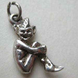 May include: A silver charm in the shape of a seated imp or goblin, with pointed ears and a mischievous grin. The charm is detailed with visible facial features and is suspended from a small loop. The imp is hugging its knees.