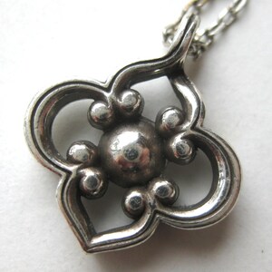 May include: A silver-toned metal pendant featuring a four-lobed, clover-like shape with a central circular sphere surrounded by four smaller spheres. The pendant is attached to a thin silver-toned metal chain link necklace.