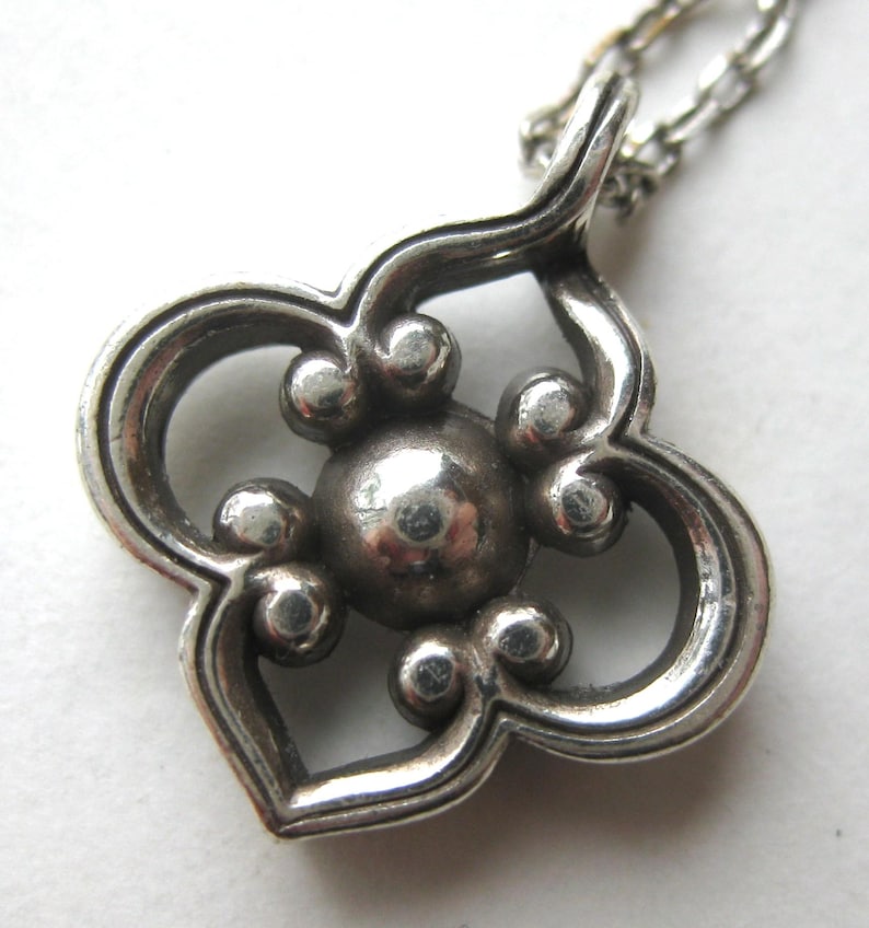 May include: A silver-toned metal pendant featuring a four-lobed, clover-like shape with a central circular sphere surrounded by four smaller spheres. The pendant is attached to a thin silver-toned metal chain link necklace.