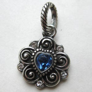 May include: A silver-toned metal pendant featuring a central heart-shaped blue crystal surrounded by swirling filigree patterns and four small clear round crystals. The piece includes a twisted rope-textured bail at the top for attachment.