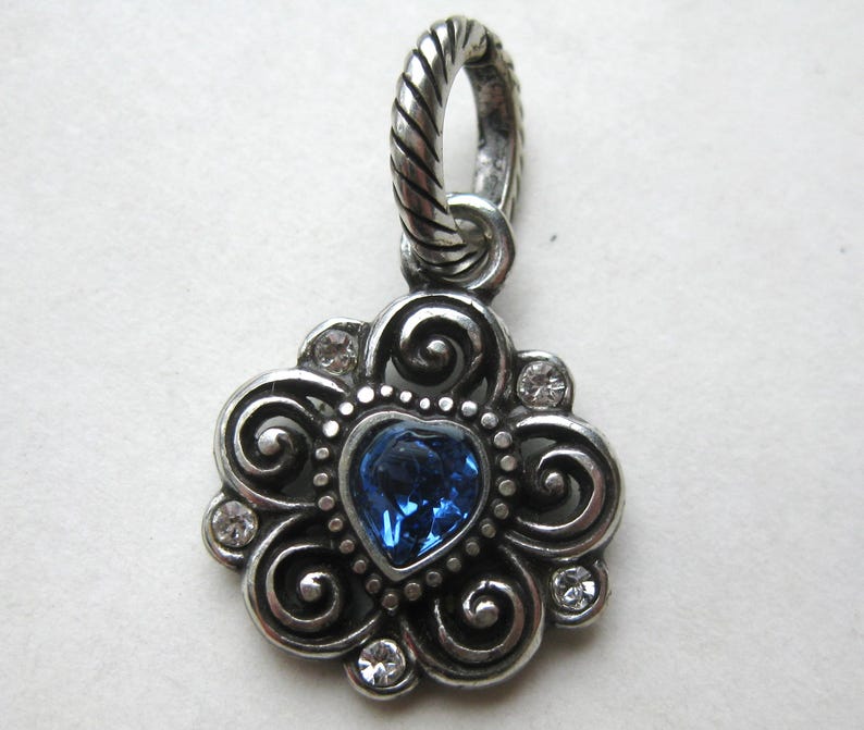 May include: A silver-toned metal pendant featuring a central heart-shaped blue crystal surrounded by swirling filigree patterns and four small clear round crystals. The piece includes a twisted rope-textured bail at the top for attachment.