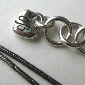 May include: Close-up of a silver necklace with a heart-shaped charm. The charm has the initials "JB" engraved on it. The necklace features a chain of interlocking rings and a thin, textured chain.