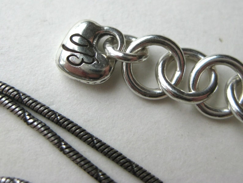May include: Close-up of a silver necklace with a heart-shaped charm. The charm has the initials "JB" engraved on it. The necklace features a chain of interlocking rings and a thin, textured chain.