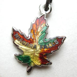 Op de afbeelding: Een kleurrijke esdoornbladbedel met de woorden "NIAGARA FALLS" in witte letters. Het blad heeft een kleurovergang van rood, oranje, geel en groen, met een zilverkleurige metalen rand en lus.