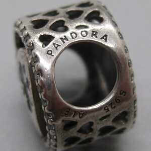 May include: A silver charm bead featuring a repeating cut-out heart pattern across its surface. The word PANDORA is engraved along the top edge of the circular opening, while the text S925 ALE is stamped along the bottom edge of the metal bead.