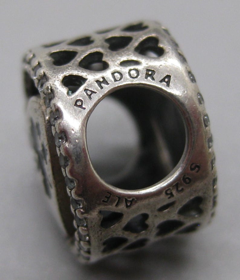 May include: A silver charm bead featuring a repeating cut-out heart pattern across its surface. The word PANDORA is engraved along the top edge of the circular opening, while the text S925 ALE is stamped along the bottom edge of the metal bead.