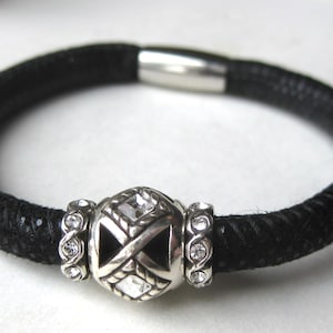 Puede incluir: Una pulsera negra texturizada con una cuenta central plateada con detalles de cristales transparentes, flanqueada por dos anillos plateados m&aacute;s peque&ntilde;os incrustados con cristales. La pulsera tiene un cierre magn&eacute;tico cil&iacute;ndrico liso.