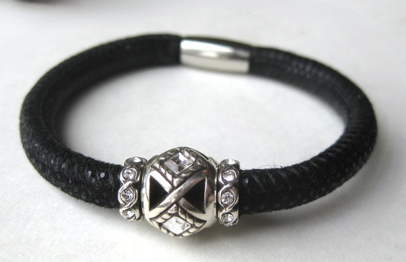 Puede incluir: Una pulsera negra texturizada con una cuenta central plateada con detalles de cristales transparentes, flanqueada por dos anillos plateados m&aacute;s peque&ntilde;os incrustados con cristales. La pulsera tiene un cierre magn&eacute;tico cil&iacute;ndrico liso.