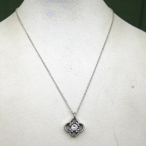 May include: A silver-toned metal necklace featuring a delicate chain and a pendant shaped like a four-lobed quatrefoil. The center of the pendant is set with a single, clear, round-cut gemstone that reflects light, creating a bright, sparkling focal point.
