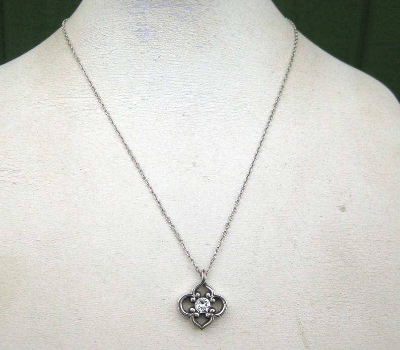 May include: A silver-toned metal necklace featuring a delicate chain and a pendant shaped like a four-lobed quatrefoil. The center of the pendant is set with a single, clear, round-cut gemstone that reflects light, creating a bright, sparkling focal point.