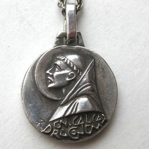 May include: A circular silver-toned metal pendant featuring a relief profile of a monk with a halo, wearing a hooded robe. The pendant is attached to a silver-toned chain. The lower portion of the pendant displays engraved text that reads CN. CALEX DRAGONUM.