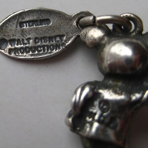 May include: A close-up of a silver-colored charm featuring a cartoon character. The charm is attached to a tag that reads "STERLING" and "WALT DISNEY PRODUCTIONS". The charm has a loop for attachment.