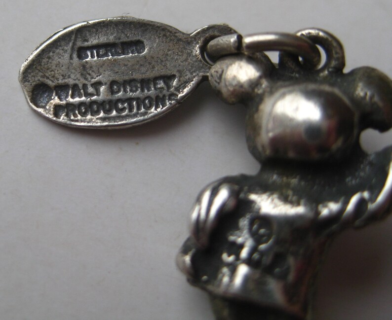 May include: A close-up of a silver-colored charm featuring a cartoon character. The charm is attached to a tag that reads "STERLING" and "WALT DISNEY PRODUCTIONS". The charm has a loop for attachment.