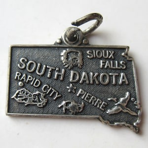 May include: A silver-colored charm in the shape of South Dakota. The charm features the state name and the cities of Rapid City, Sioux Falls, and Pierre. Small embossed images of a bison, a bird, and a mountain range are also visible.
