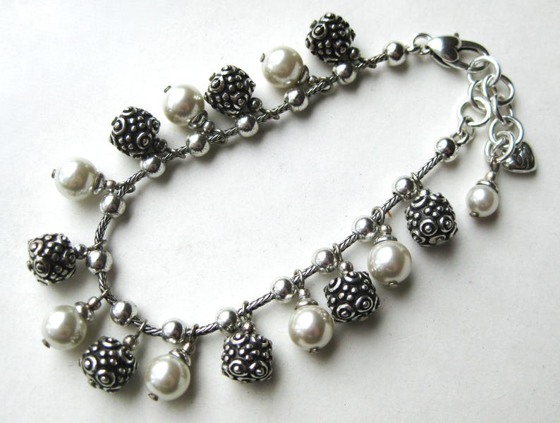 Puede incluir: Pulsera plateada con perlas de imitaci&oacute;n y cuentas decorativas texturizadas. La pulsera tiene un cierre de mosquet&oacute;n y un peque&ntilde;o dije de coraz&oacute;n. Las cuentas y perlas cuelgan de la cadena.