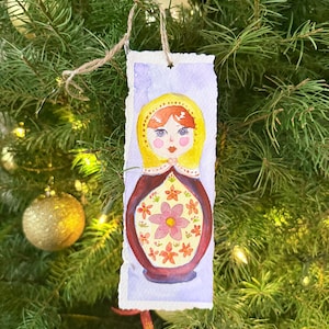 Puede incluir: Adorno navideño de acuarela con un diseño de muñeca Matryoshka. La muñeca tiene un pañuelo amarillo, pelo rojo y un diseño floral en un cuerpo granate. El adorno rectangular tiene un borde rasgado y un colgador de cuerda.