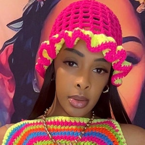 May include: A bright pink crochet hat with a yellow trim. The hat is worn with a rainbow-colored crochet top. The image is a close-up shot of a person wearing the hat and top.