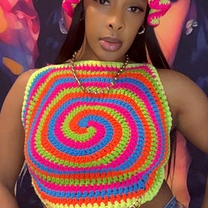 May include: A vibrant, handmade crochet top and matching hat. The top features a spiral design in neon colors: pink, green, blue, and orange. The hat is pink with a yellow trim. A gold chain belt adds a touch of glamour.