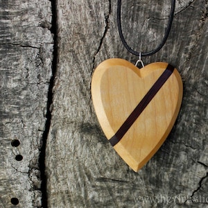 May include: Wooden heart pendant with a dark purple stripe across the front. The heart-shaped pendant is light brown and hangs from a black cord. The background is a weathered, gray wooden surface.