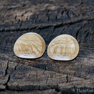 May include: Two wooden stud earrings with a light brown, wood-grain pattern. The earrings are oval-shaped and sit on a dark, textured wooden surface. The earrings are a natural, neutral color.