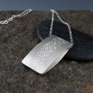 May include: A silver necklace with a rectangular pendant featuring a textured design of small circles. The pendant is hanging on a silver chain.