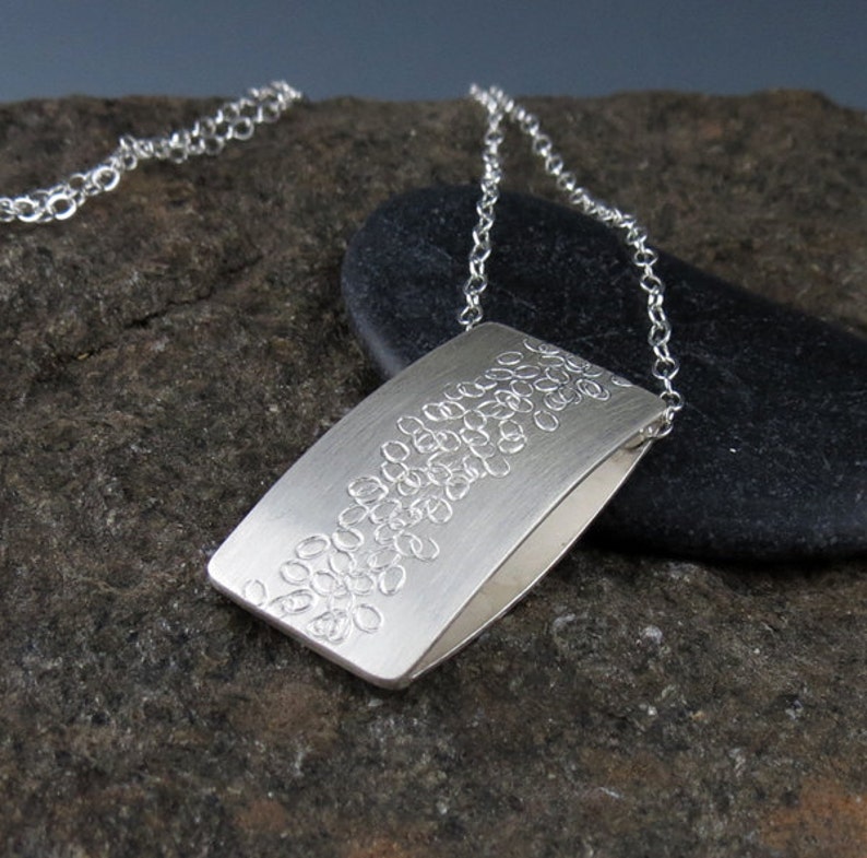 May include: A silver necklace with a rectangular pendant featuring a textured design of small circles. The pendant is hanging on a silver chain.