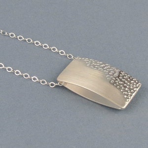 May include: A silver necklace with a rectangular pendant. The pendant has a smooth, brushed finish on one side and a textured, hammered finish on the other side.