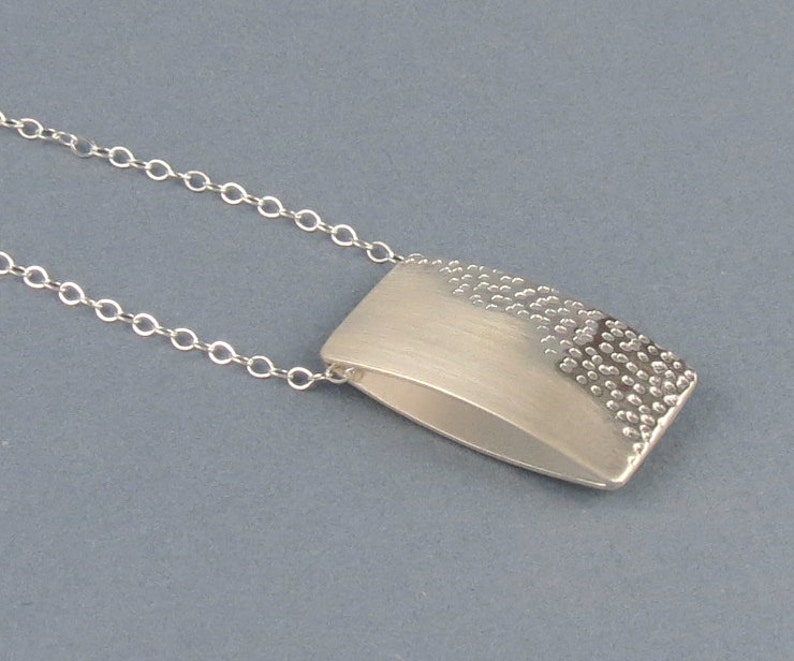 May include: A silver necklace with a rectangular pendant. The pendant has a smooth, brushed finish on one side and a textured, hammered finish on the other side.