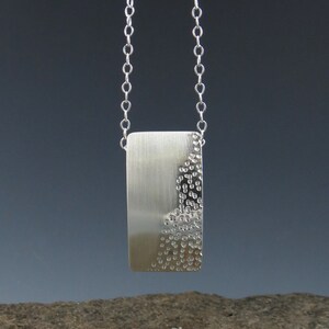 May include: A silver necklace with a rectangular pendant. The pendant has a smooth, brushed finish on one side and a textured, dotted pattern on the other side.