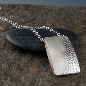 May include: A silver necklace with a rectangular pendant. The pendant has a smooth, brushed finish on one side and a textured, dotted pattern on the other side.