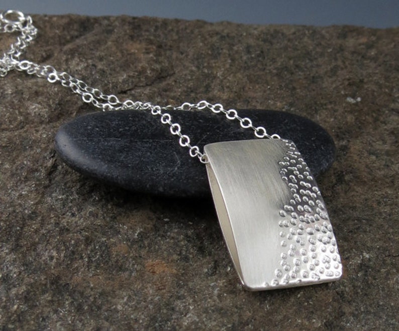 May include: A silver necklace with a rectangular pendant. The pendant has a smooth, brushed finish on one side and a textured, dotted pattern on the other side.